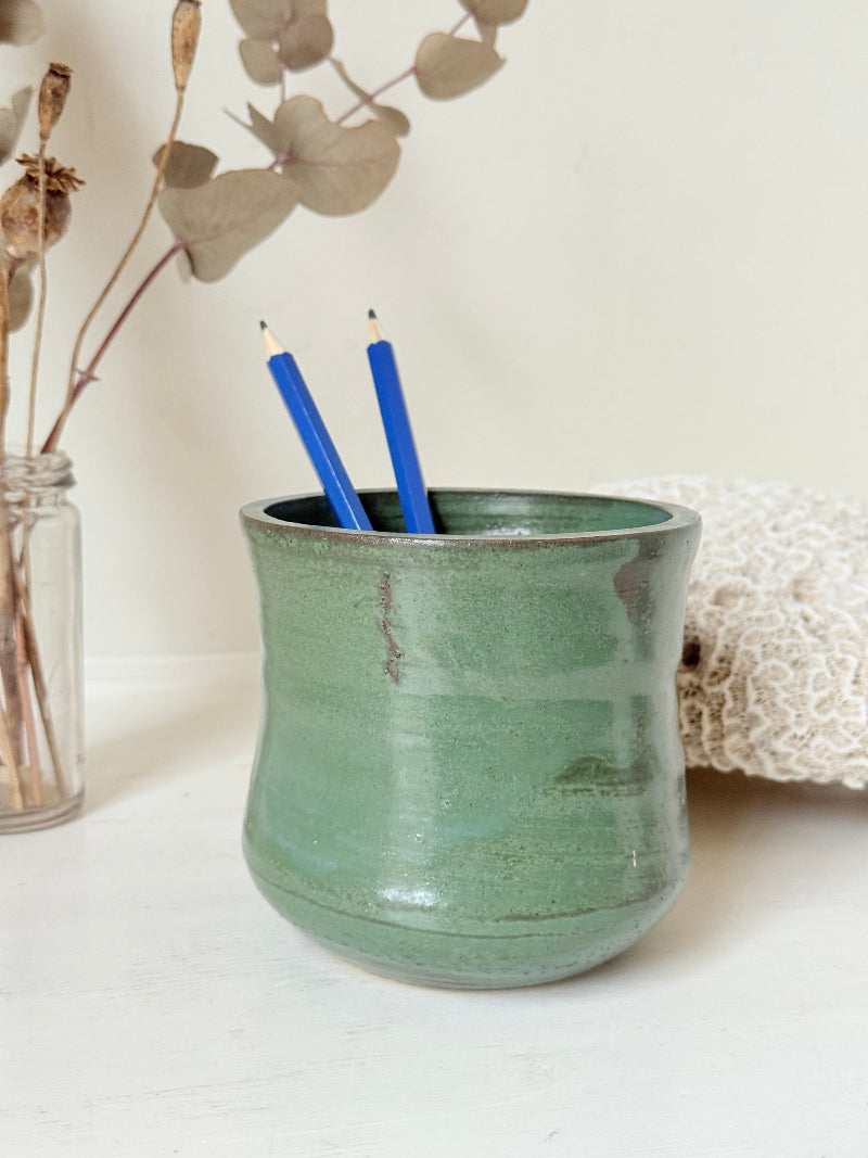 Studio pottery pencil pot in green glaze, handmade on the wheel, photographed on a white surface with pencils inside.