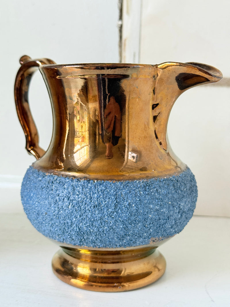A vintage-style metallic pitcher with a reflective gold finish and a textured blue lower section, shown on a white surface against a white background.