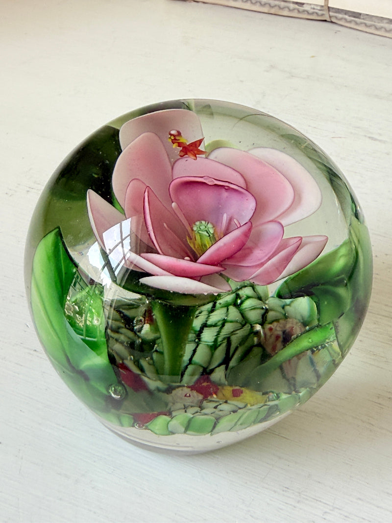 Large clear glass paperweight with a pink flower and green leaves inside, photographed on a white surface