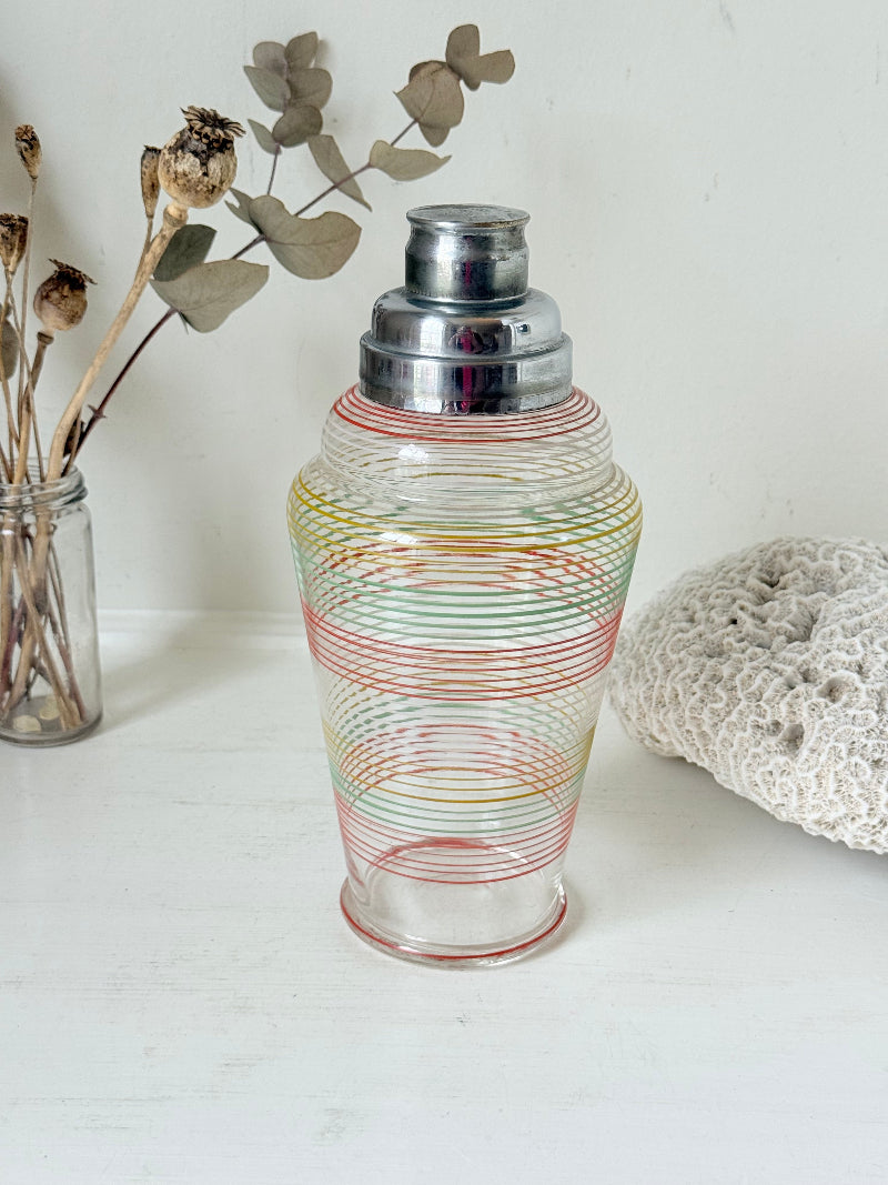 Vintage glass cocktail shaker with multicoloured stripes and silver lid photographed on a white surface.