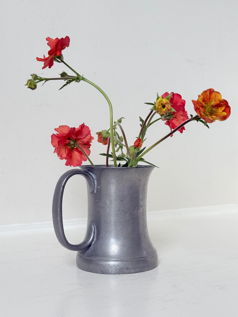 vintage pewter tankard with crown and rose mark and handle, styled with red and orange flowers on white surface