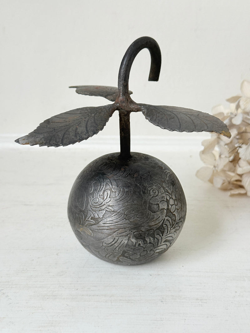 A vintage-style metal hanging ornament shaped like a round engraved sphere topped with a curved hook and leaf-like wings, displayed against a light neutral background with dried flowers at the side.