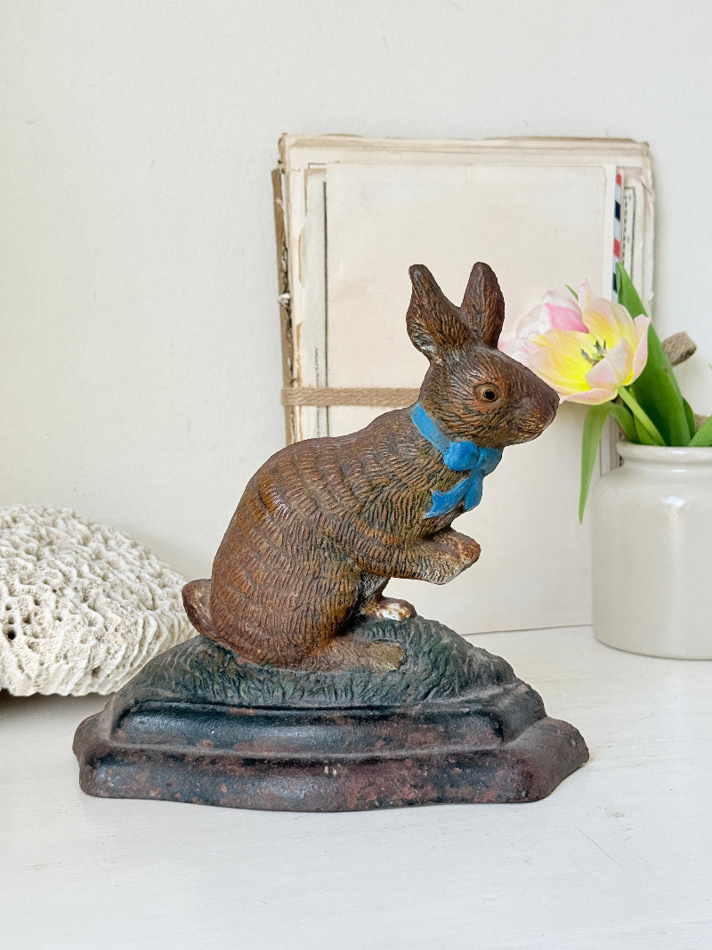 A vintage-style metal rabbit figurine with a textured brown finish and a bright blue bow tie, sitting on a dark base. The figurine is styled on a white surface with a white ceramic vase containing pink tulips and a rustic book in the background.