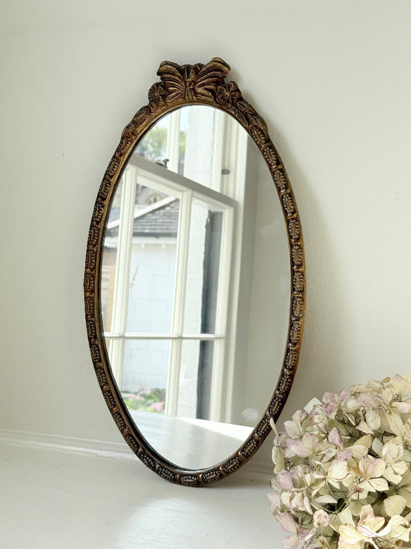 Vintage oval wall mirror with decorative gilt frame and bow crest, styled against a neutral wall with hydrangeas.