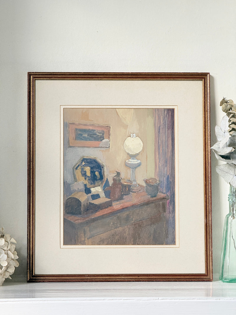 A framed art print depicting a still life interior scene with a table, lamp, and blue-and-white vase, displayed on a white surface against a light wall.