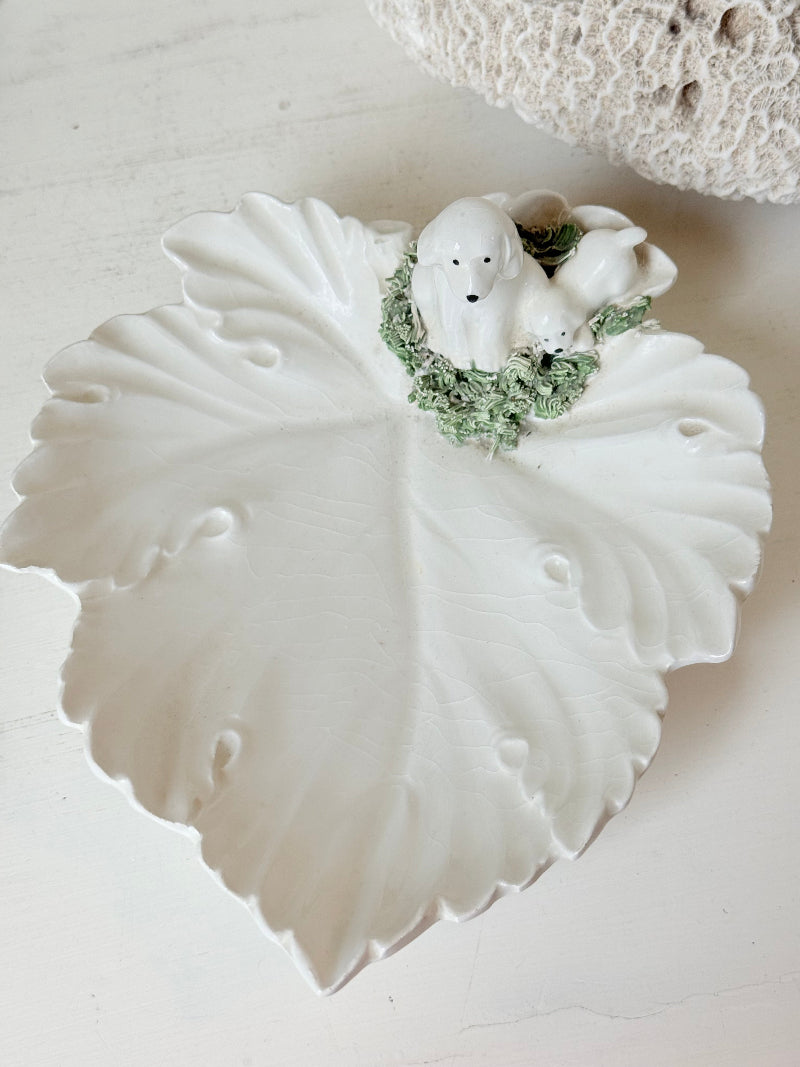 Italian ceramic dish featuring two white dogs on a sculpted leaf base, photographed on a white surface.