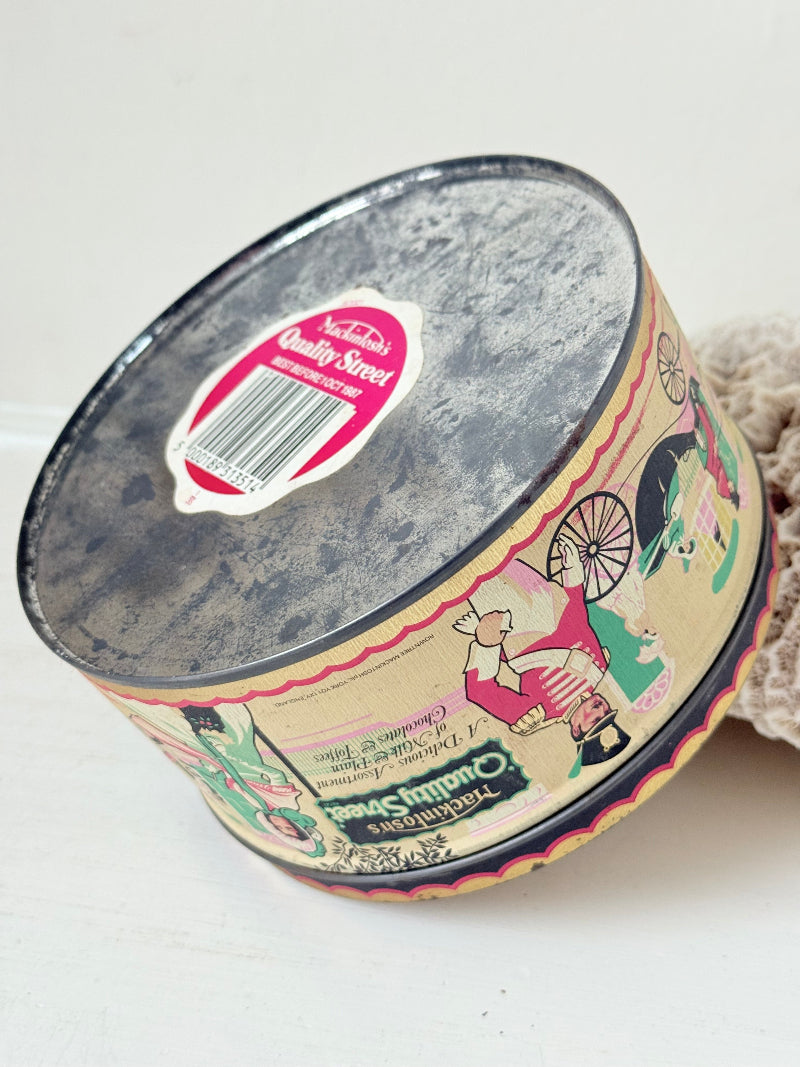 Vintage Quality Street tin from 1986 with colourful illustrated street scenes and pastel design, photographed on a white surface.