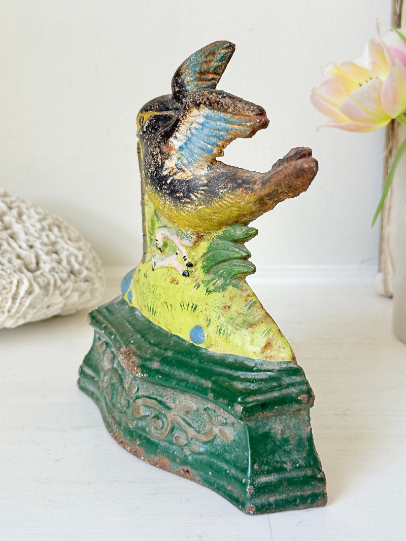 A vintage hand-painted ceramic bird figurine on an ornate green base, displayed on a white surface with soft-focus decorative props in the background.