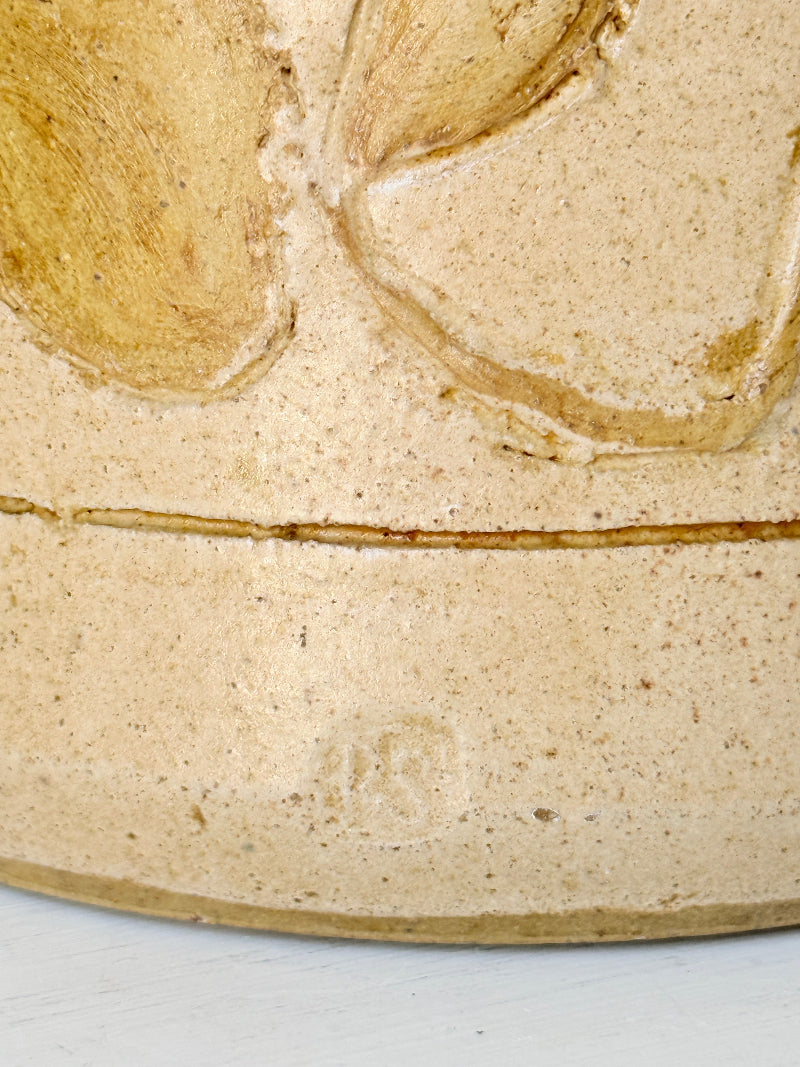 Close-up of a beige ceramic surface with a raised floral relief and a horizontal seam, showing a speckled clay texture and a faint stamped mark near the bottom.