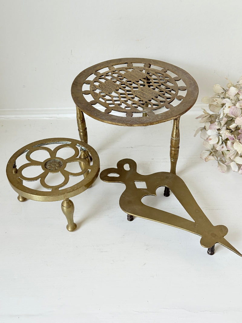 Vintage brass trivet trio with ornate cutwork designs and aged patina, photographed on a white surface