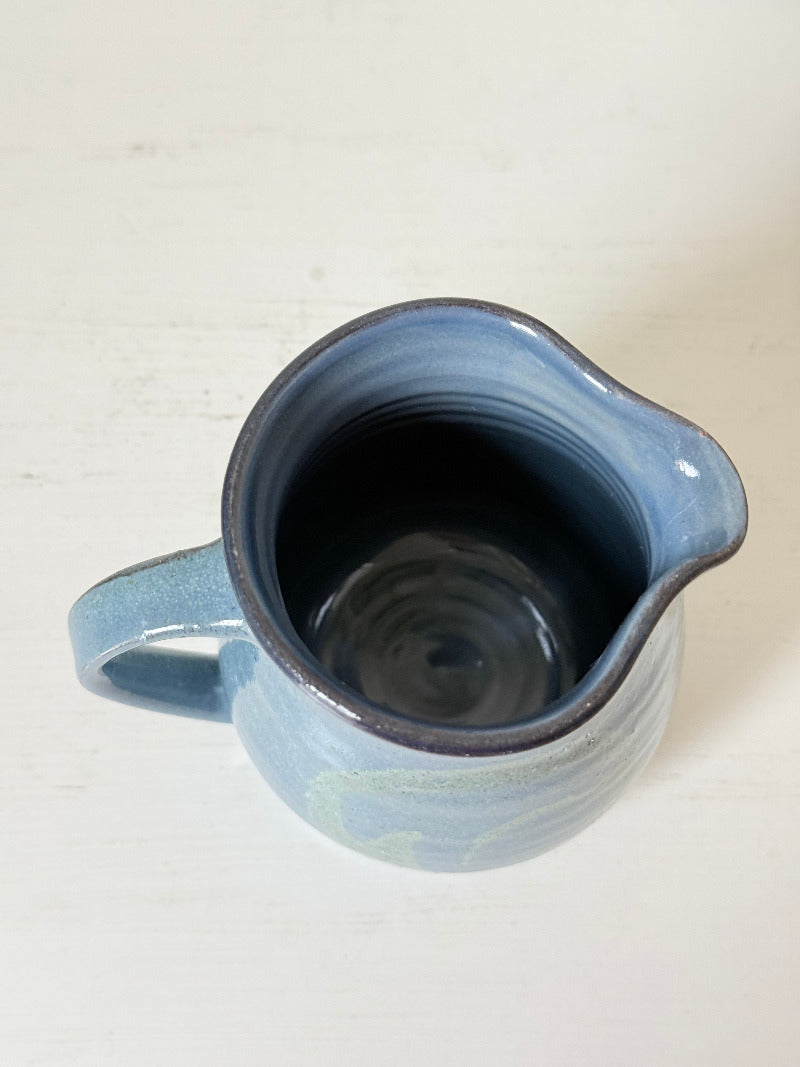 Vintage studio pottery milk jug with blue and green glaze, handmade with a rounded form and sturdy handle, styled on a white surface.