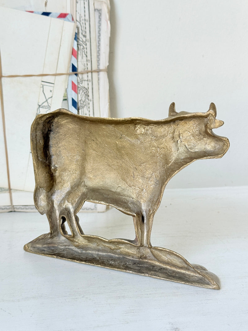 A rustic, hand-painted metallic cow figurine in a distressed gold/bronze finish, standing on a matching base against a white background with vintage mail in the soft-focus background.