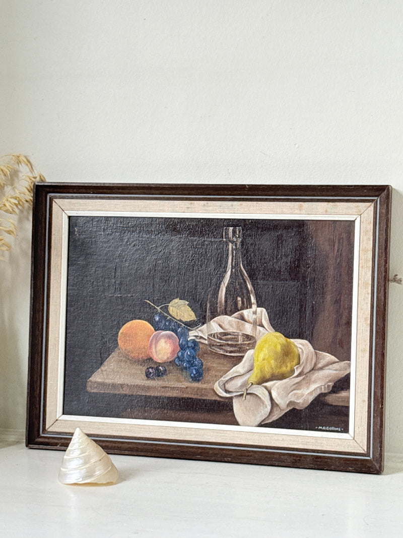 A framed still life oil painting depicting fruit, grapes, a pear, and a glass bottle on a tablecloth, set in a dark brown wooden frame with a cream mat, leaning against a light wall.