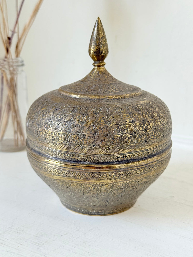 Indian Benares brass lidded jar with pointed finial and darkened interior, photographed on a white surface