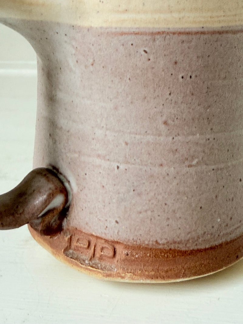 Studio pottery milk jug in cream and lavender glaze with impressed initials, photographed on a white surface.