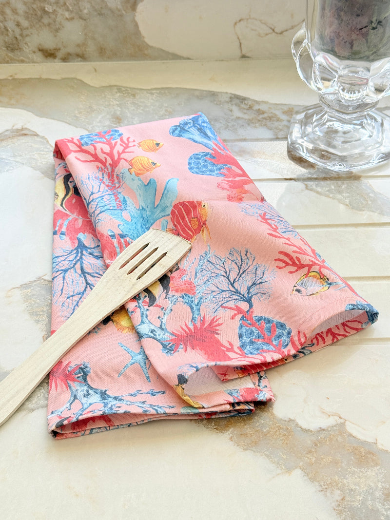 Warm peach coral and tropical fish print cotton tea towel with wooden forks on a marble surface beside cut glass