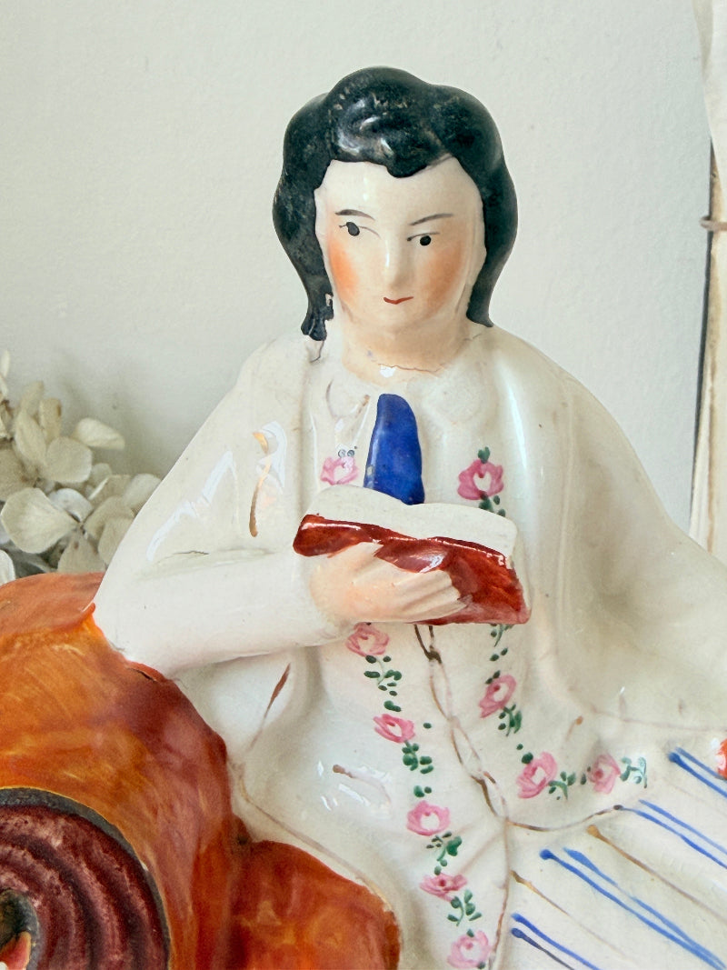 A vintage ceramic figurine of a man with black hair, wearing a white jacket decorated with pink floral patterns, holding a red book and a blue tie, set against a light background.