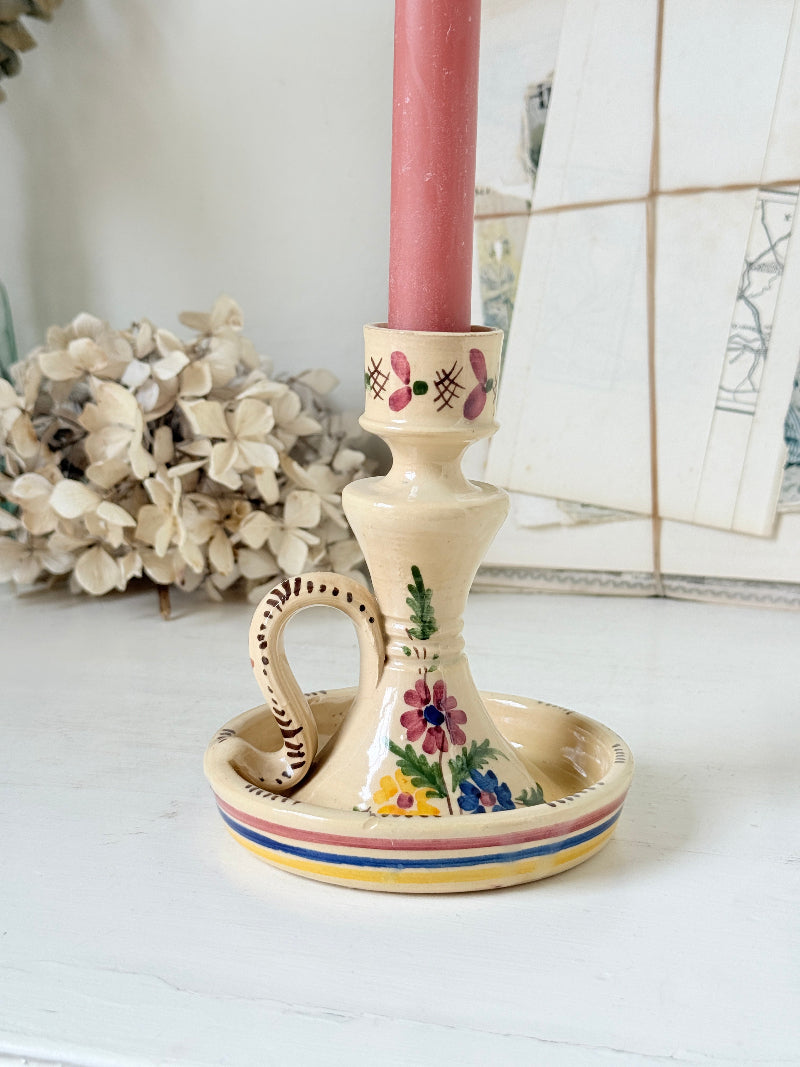 A vintage-style ceramic candlestick holder with a handle, a matching saucer, and a pink candle inserted, decorated with colorful floral and geometric patterns, set on a white surface with dried flowers in the background.