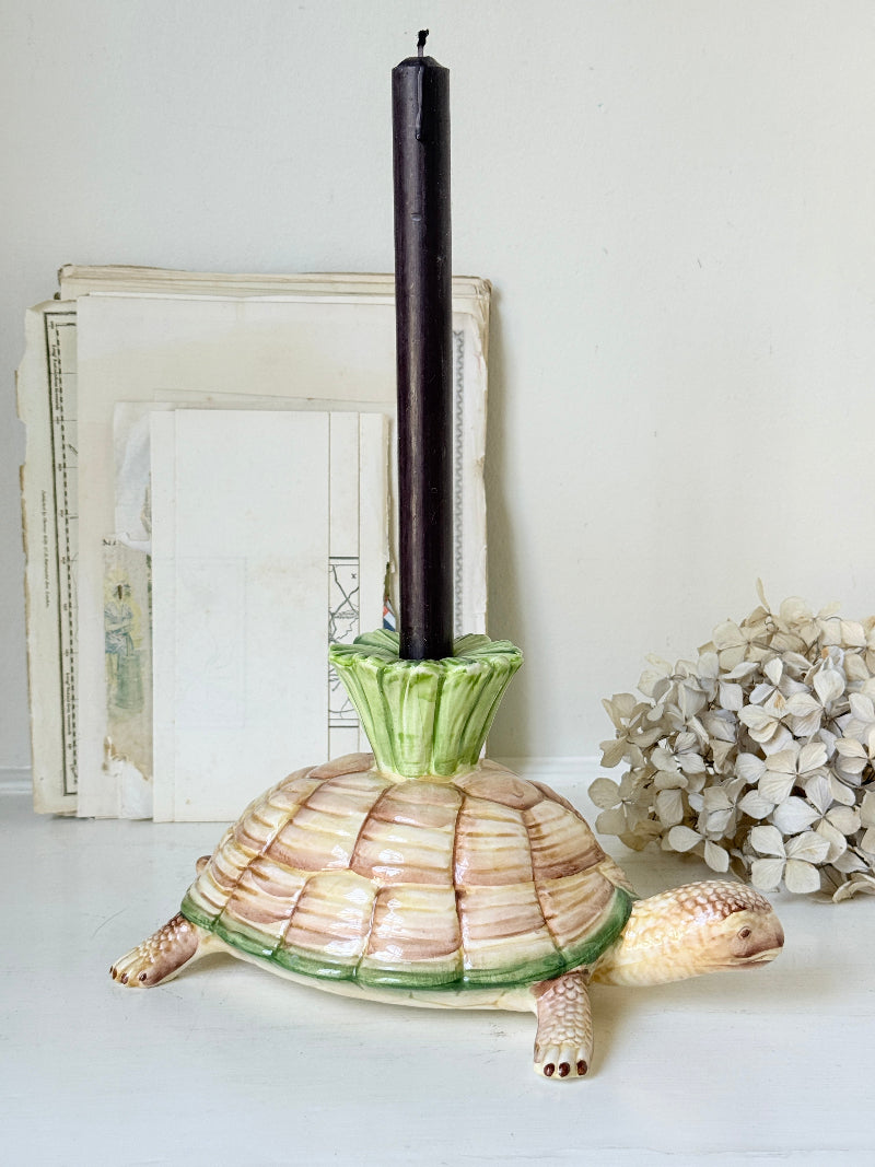 A ceramic tortoise candle holder with a cream and green shell, holding a tall black taper candle, styled on a white surface with vintage papers and dried flowers in the background.