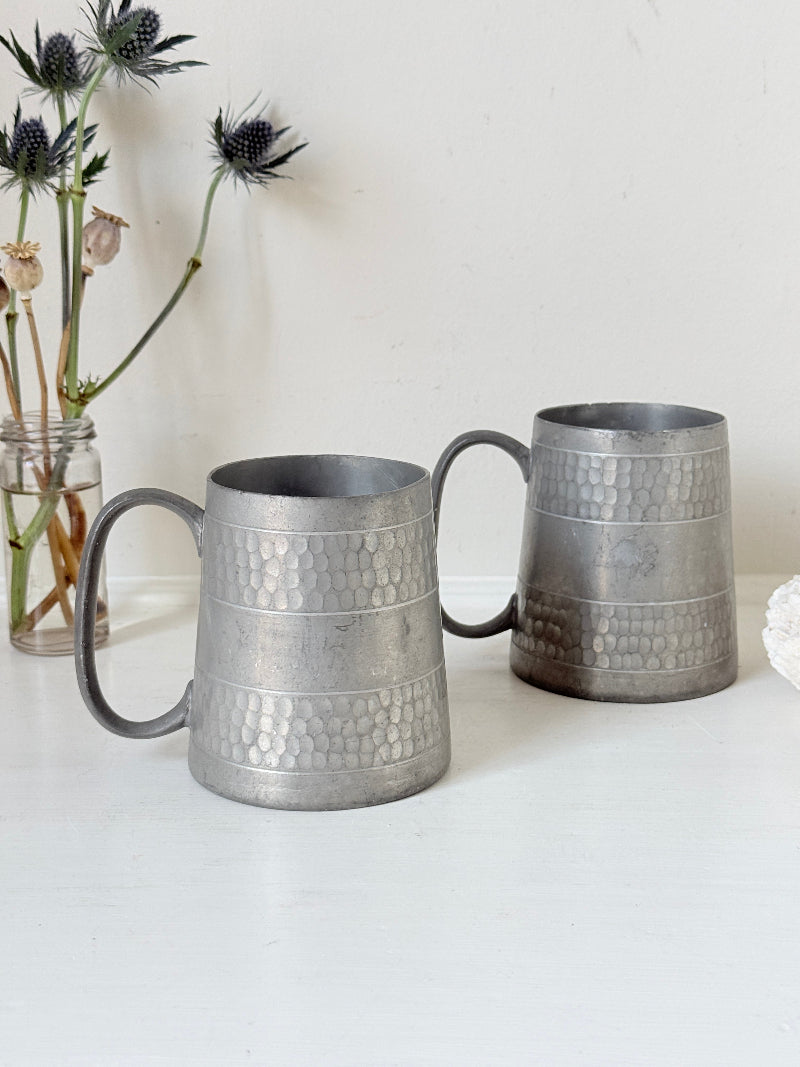 Pair of vintage pewter tankards with handles and textured detail, photographed on a white surface