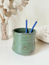 Studio pottery pencil pot in green glaze, handmade on the wheel, photographed on a white surface with pencils inside.
