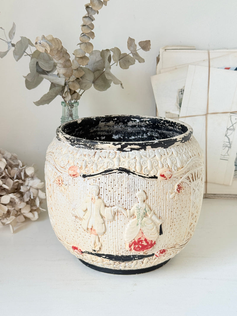 A vintage-style ceramic planter pot with raised figures and floral details, featuring a distressed cream finish with black and red accents, styled on a white surface with dried flowers and papers in the background.