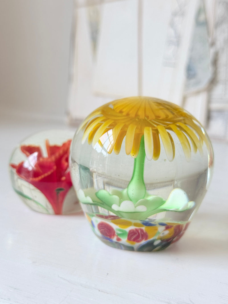 Pair of yellow and red flower glass paperweights with green stems, photographed on a white surface