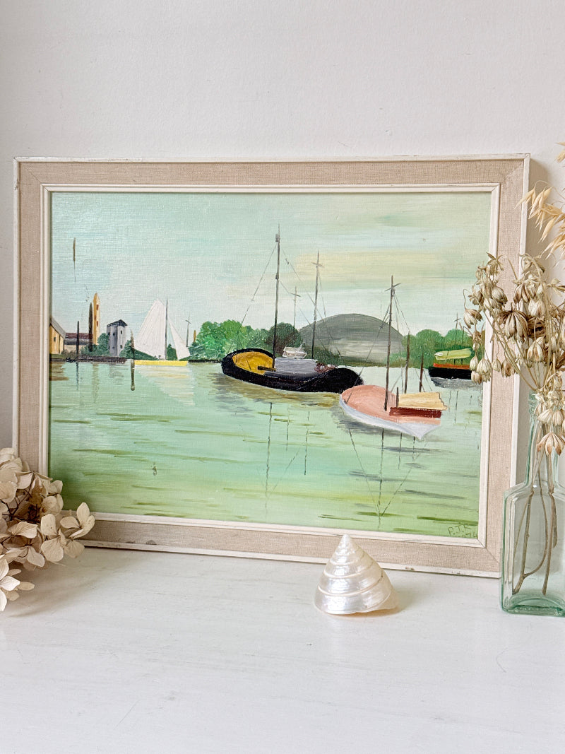 A framed watercolor painting of a harbor scene with boats on greenish water, displayed on a white surface with dried flowers and a shell as props.