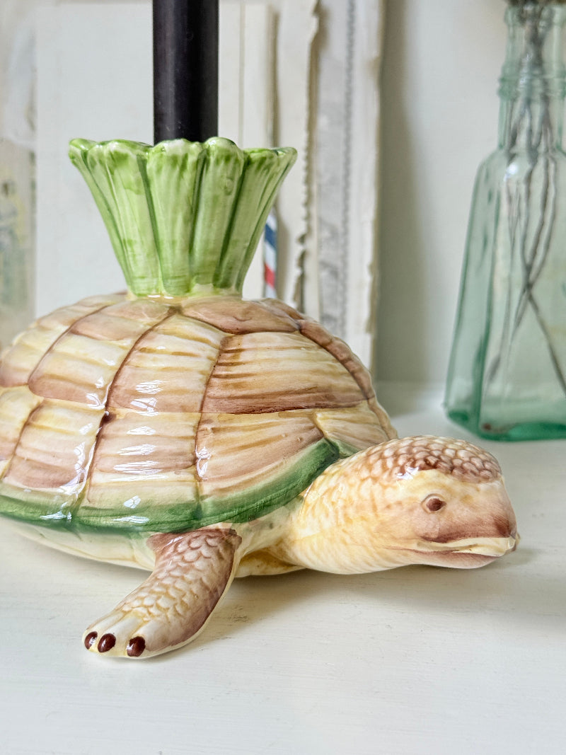 A ceramic turtle-shaped candle holder with a glossy finish, featuring a beige and brown shell, a green flared top, and a cream-colored head and front legs, shown on a tabletop with a glass bottle in the background.