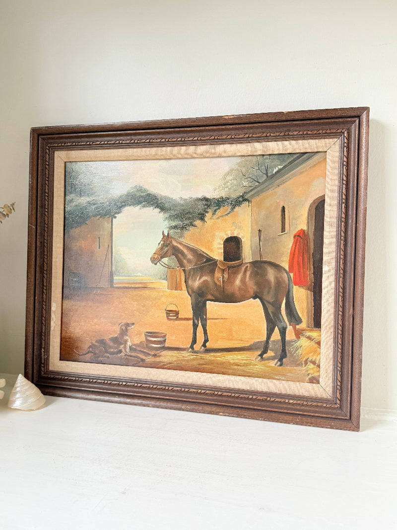 A framed vintage oil painting depicting a brown horse standing in a stable courtyard, set in a dark brown wooden frame on a white surface against a light beige wall.