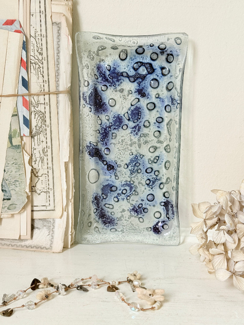 A rectangular glass dish with a textured, bubbly pattern in shades of blue, purple, and white, styled on a white surface next to vintage letters and dried flowers.