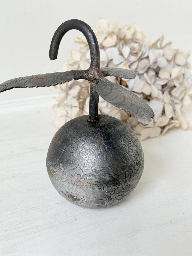 A vintage-style metal hanging ornament with a round engraved ball base, a curved hook top, and leaf-like wings extending from the sides, shown on a white surface with dried flowers in the background.