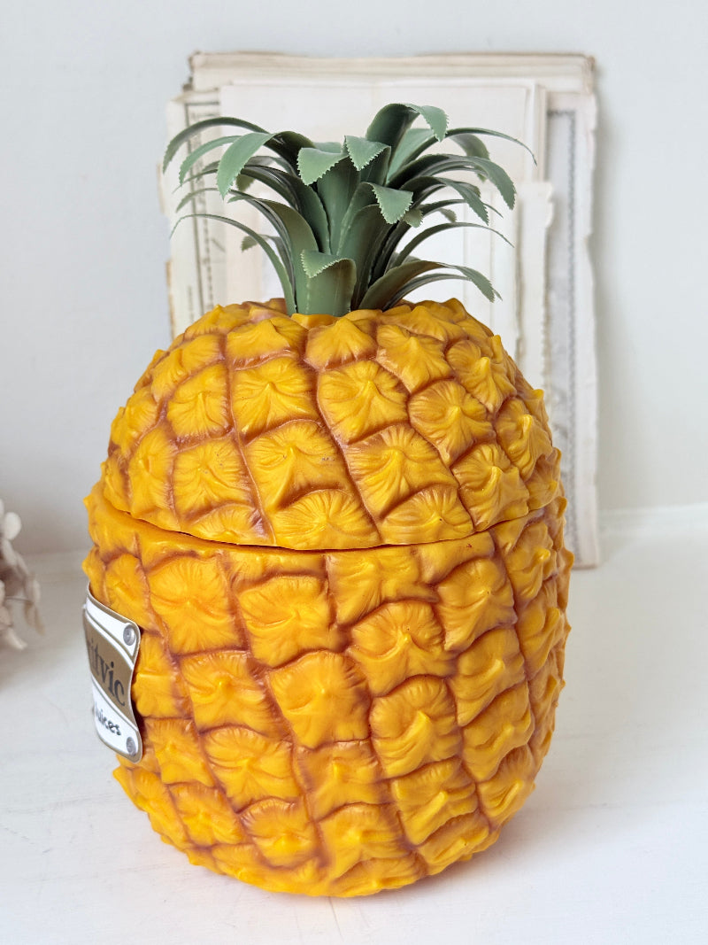 A pineapple-shaped ceramic container with a textured yellow and orange body and green leafy top, shown on a white surface with a rustic white frame in the background.