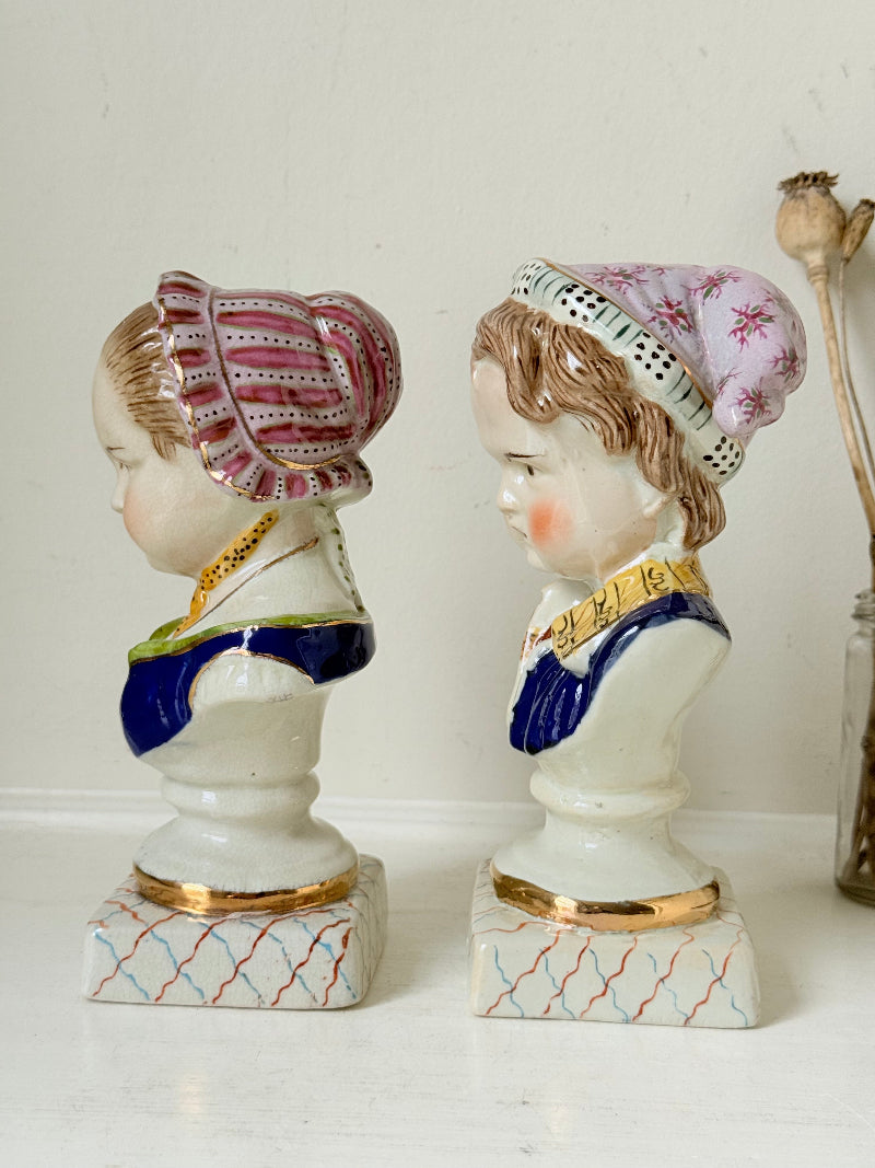 Hand painted Victorian Staffordshire ceramic children busts on white surface, photographed beside dried stems.