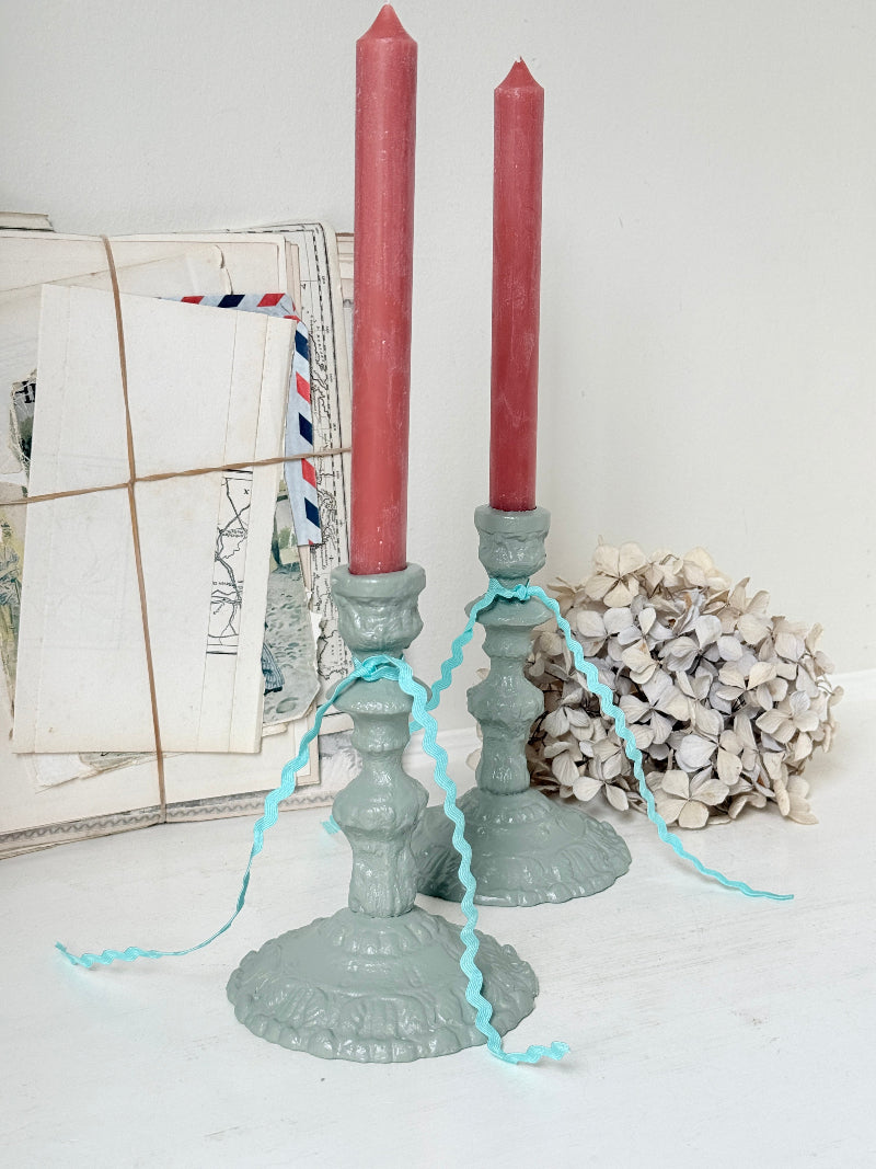 Two distressed sage green candlestick holders with red taper candles, decorated with light blue curly ribbon, styled with vintage postcards and a dried flower on a white surface.
