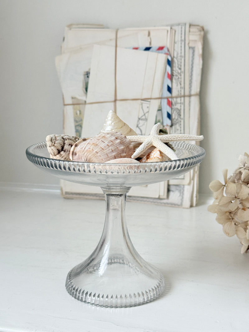 A clear glass pedestal plate filled with seashells and a starfish, styled on a white surface with vintage postcards in the background.