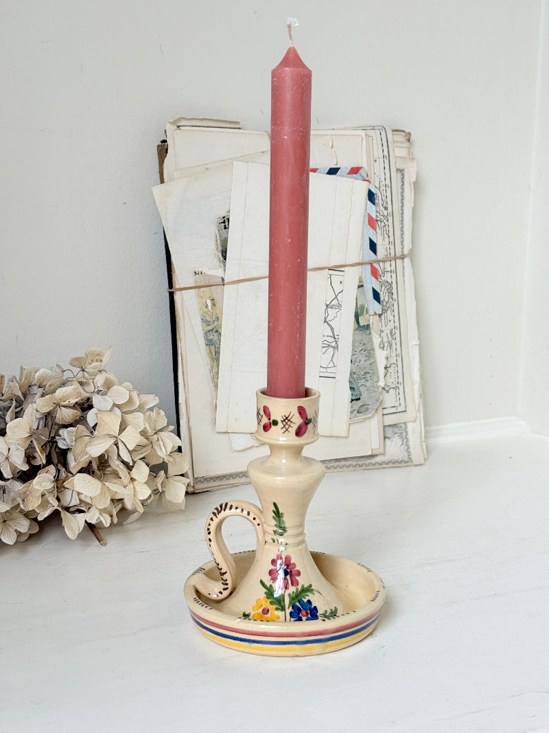 A tall dusty pink taper candle displayed in a vintage-style cream ceramic candlestick holder with floral details and a small side handle, set against a white background with old papers and dried flowers as props.