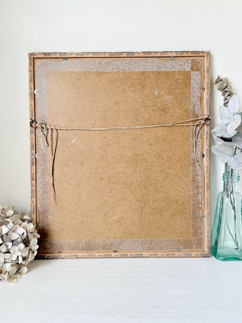 The back of a rustic wooden picture frame leaning against a white wall, with a cardboard backing secured by twine, styled with dried flowers on a white surface.