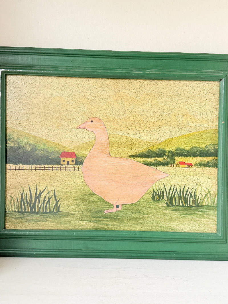 A framed folk art piece featuring a pink goose standing in a green field with a yellow sky and distant hills, enclosed in a dark green wooden frame.