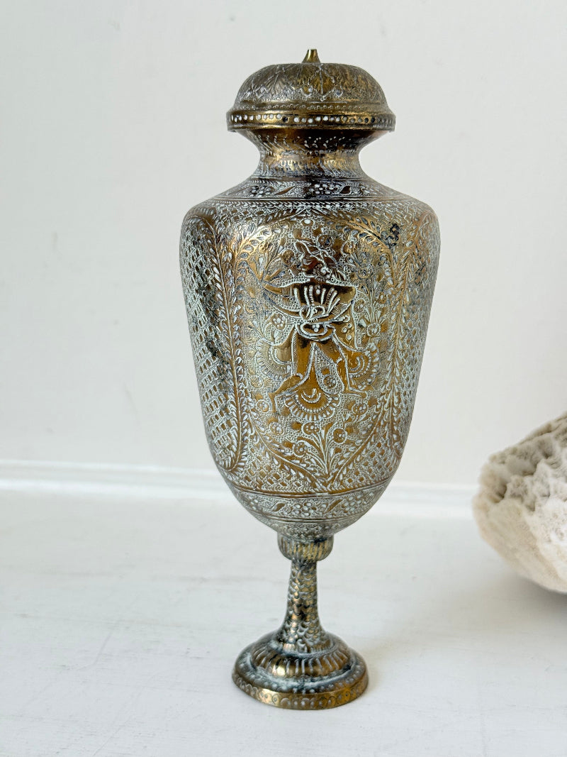 Indian Benares brass lidded vase with engraved figures and pedestal base, photographed on a white surface
