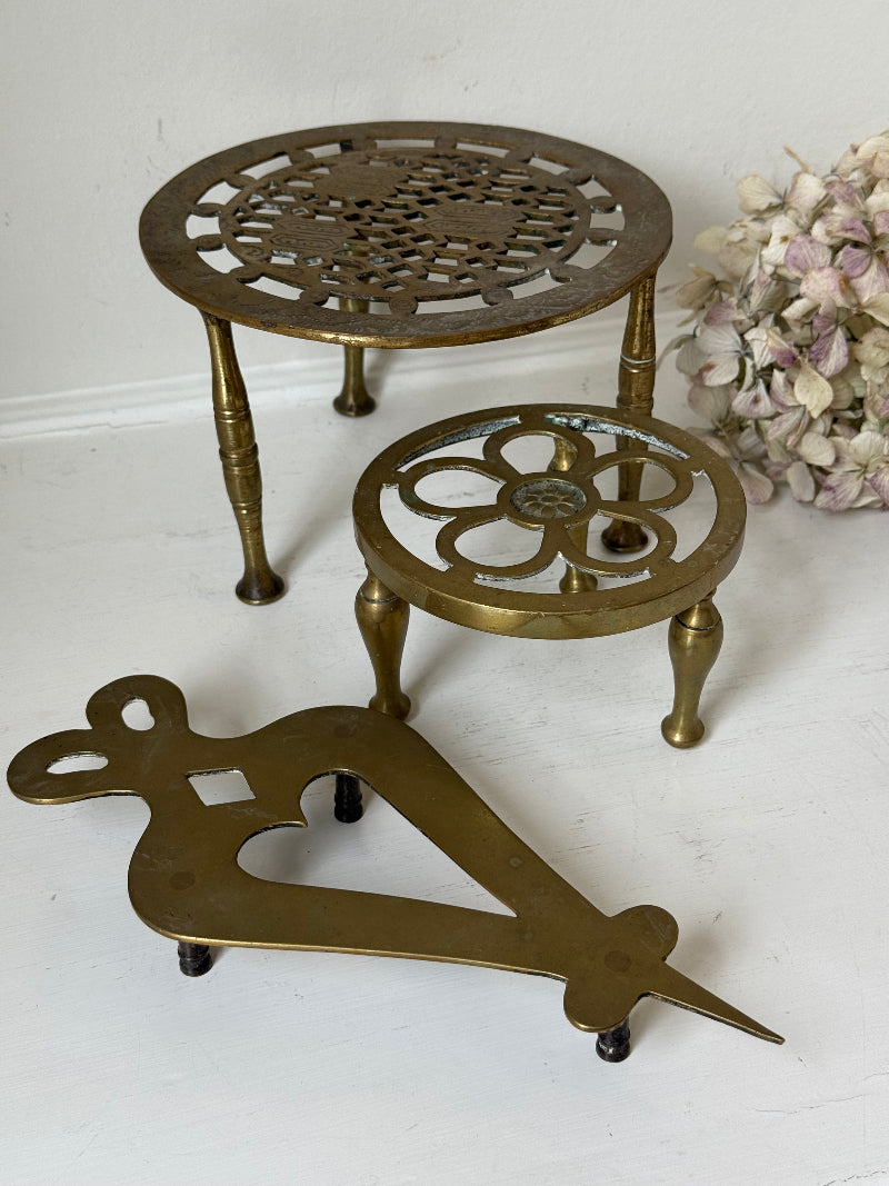 Vintage brass trivet trio with ornate cutwork designs and aged patina, photographed on a white surface
