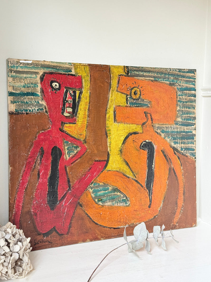 An abstract canvas painting displayed on a white surface against a white wall, featuring bold brushstrokes in red, orange, yellow, brown, and teal. Decorative white dried flowers and a textured white object sit at the base of the canvas.
