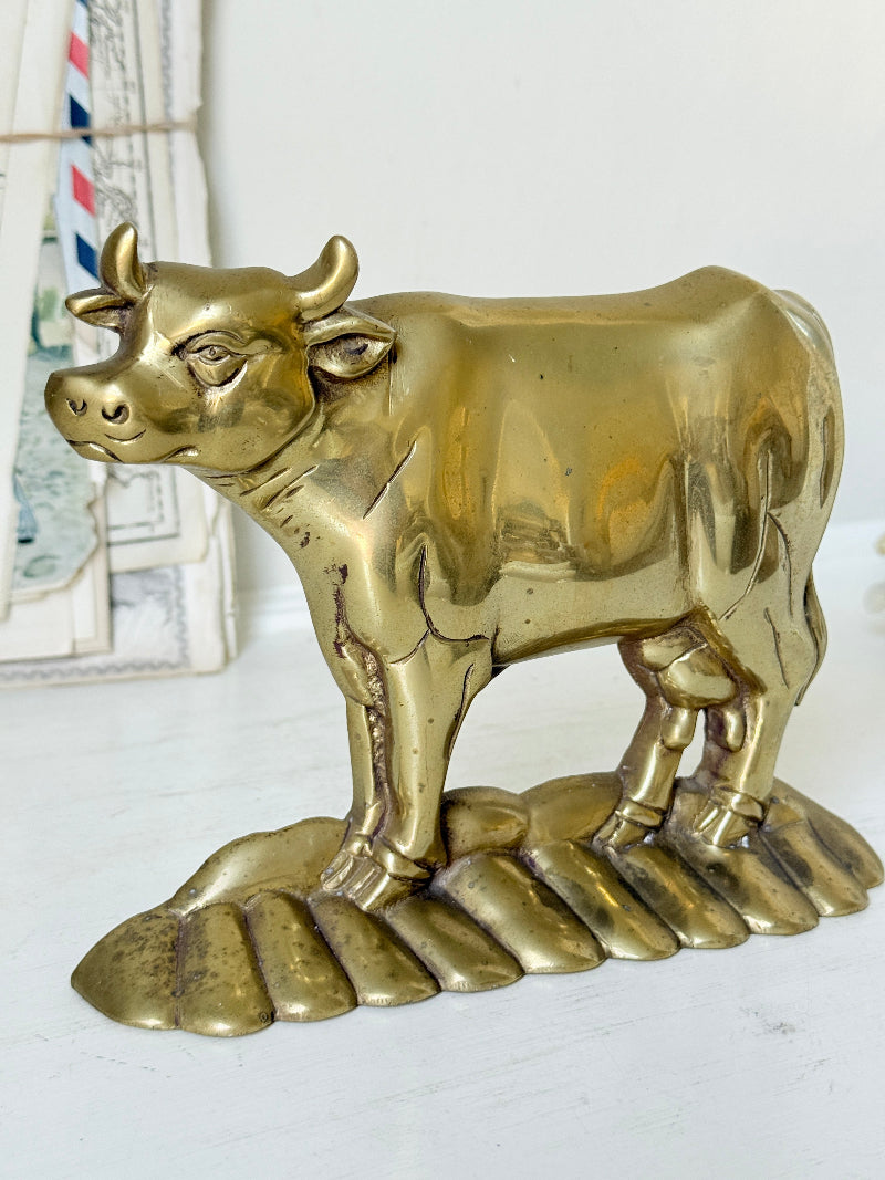 A polished brass bull figurine standing on a matching bull-hide base, photographed against a light neutral background with some vintage mail items partially visible on the left.
