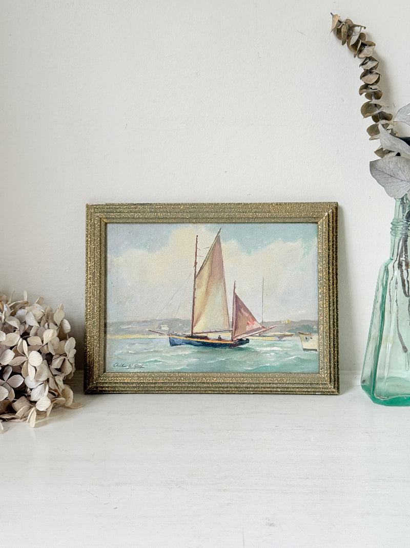 A framed vintage watercolor painting of a sailboat on the sea, displayed on a white surface with dried botanical decorations on both sides.