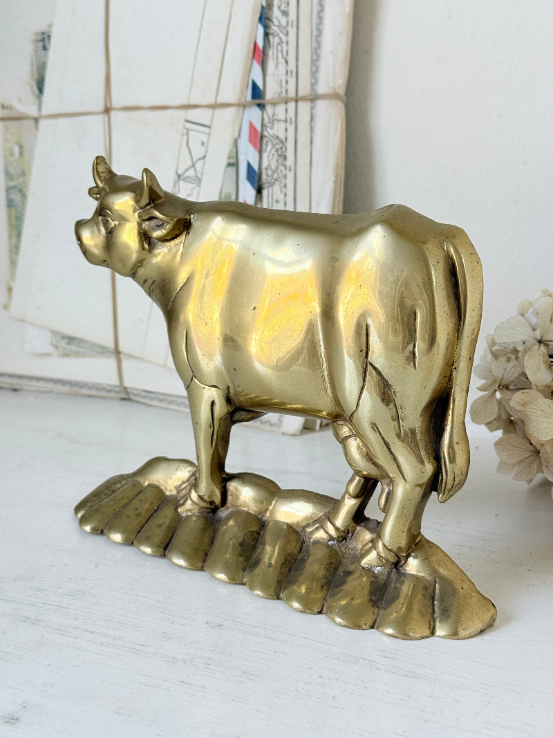 A polished brass cow figurine standing on a matching textured base, displayed on a white surface with a vintage window and dried flowers in the background.