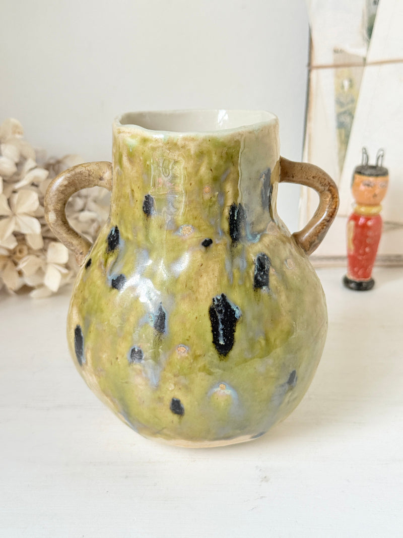 A handmade ceramic vase with a rounded body, a narrow neck, and two loop handles. The glaze has a mottled olive green, beige, and white base with irregular black speckles, set against a softly blurred background with a small figurine.