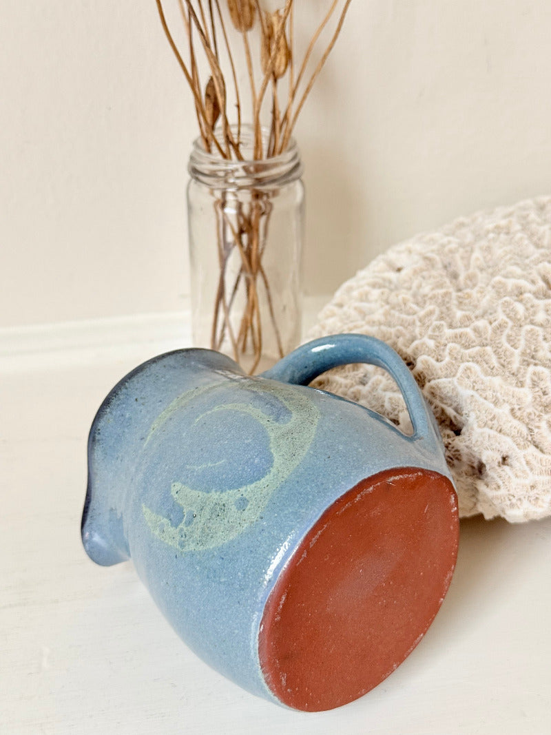 Vintage studio pottery milk jug with blue and green glaze, handmade with a rounded form and sturdy handle, styled on a white surface.