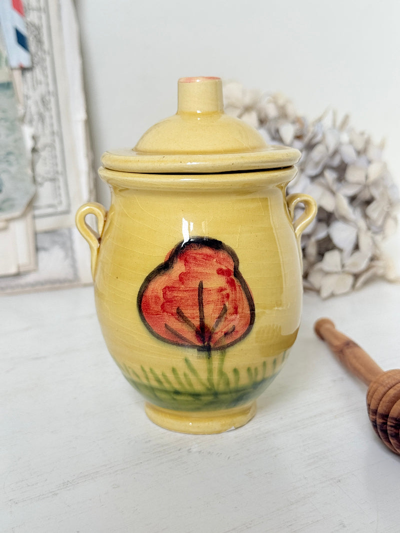 A handmade yellow ceramic lidded jar with side handles, featuring a hand-painted red poppy flower and green grass design on the front, set on a white wooden surface with blurred decorative props in the background.