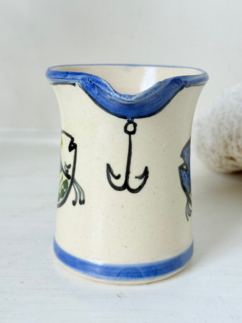 Vintage studio pottery jug with hand painted blue and green fish design on cream clay, photographed on a white surface.