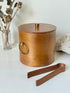 Mid century bent plywood icebox with brass handle and matching wooden tongs, styled on a white surface with dried foliage.